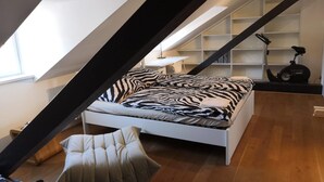 Ein modernes Schlafzimmer im Loftsstil mit schrägverdachter Decke, einem weißen Bett mit Zebra-Muster Bettwäsche, einem kleinen Hocker und einem Laufband in der Ecke.