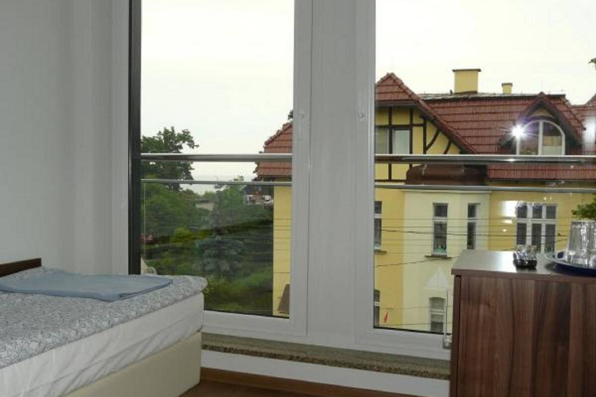 Un dormitorio con una cama, un aparador de madera y un amplio ventanal con vistas a edificios vecinos.