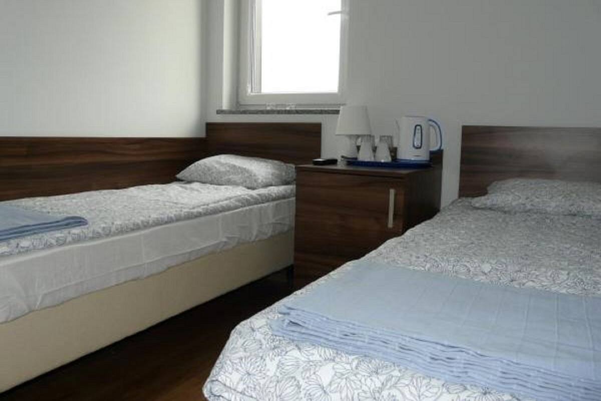 Una habitación con dos camas individuales, un cabecero de madera, una mesita de noche con lámpara y hervidor, y una ventana que deja entrar luz natural.
