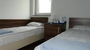 Una habitación con dos camas individuales, un cabecero de madera, una mesita de noche con lámpara y hervidor, y una ventana que deja entrar luz natural.