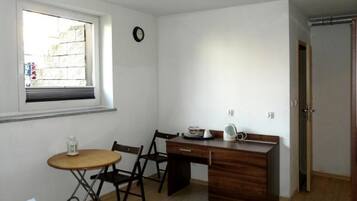 Una habitación pequeña y ordenada con un escritorio de madera, una silla, una mesita con lámpara y una ventana con persianas.