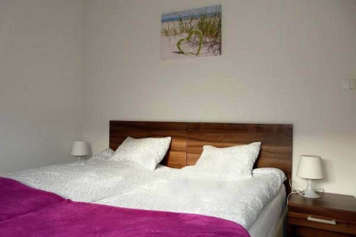 Una cama bien hecha con ropa blanca y una manta morada, un cabecero de madera, dos lámparas de noche y un pequeño cuadro de una playa en la pared.