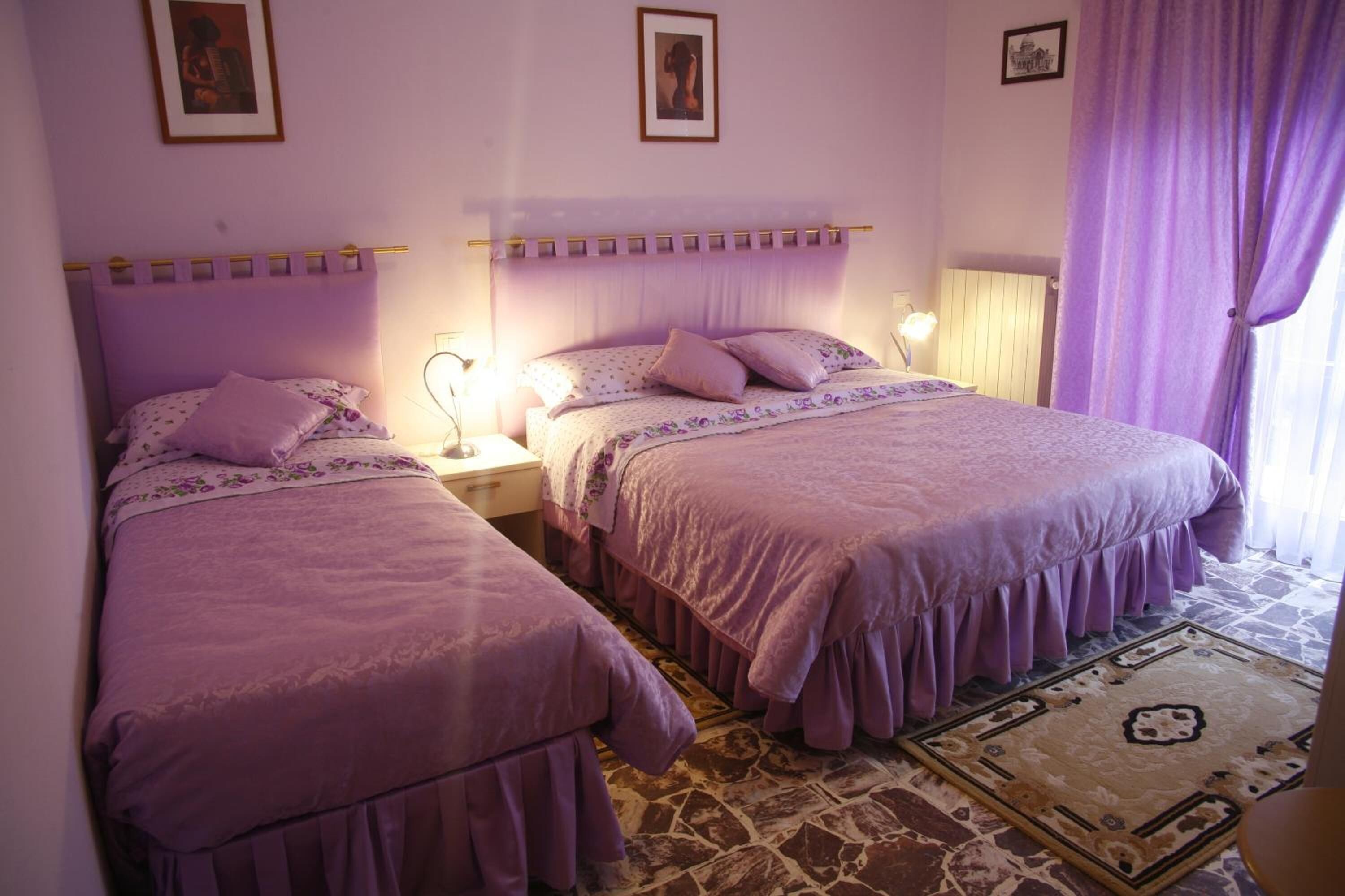A bedroom with two beds, each with a purple bedspread and matching pillows. There are two bedside tables with lamps, a patterned rug on the floor, and a window with purple curtains.