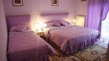 A bedroom with two beds, each with a purple bedspread and matching pillows. There are two bedside tables with lamps, a patterned rug on the floor, and a window with purple curtains.