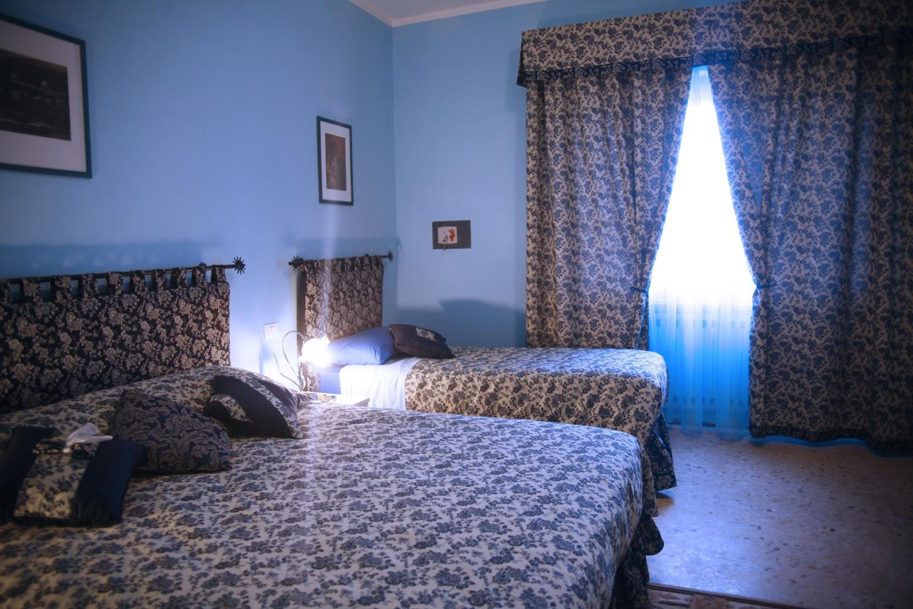 A room with two beds, floral-patterned bedding, a framed picture on the wall, and a window with patterned curtains.