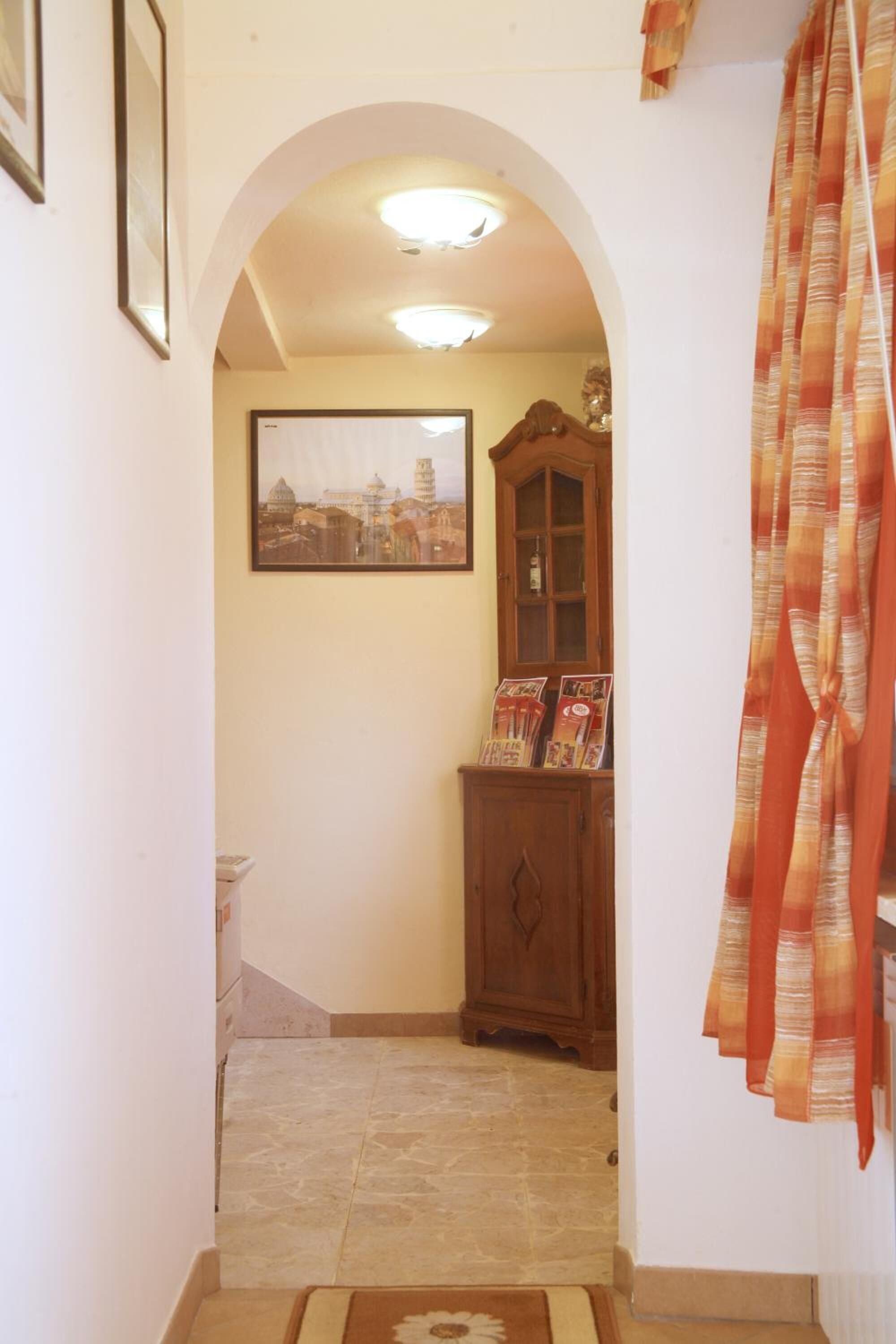 A narrow hallway with a tiled floor, a wooden cabinet with magazines, and a framed cityscape picture on the wall.