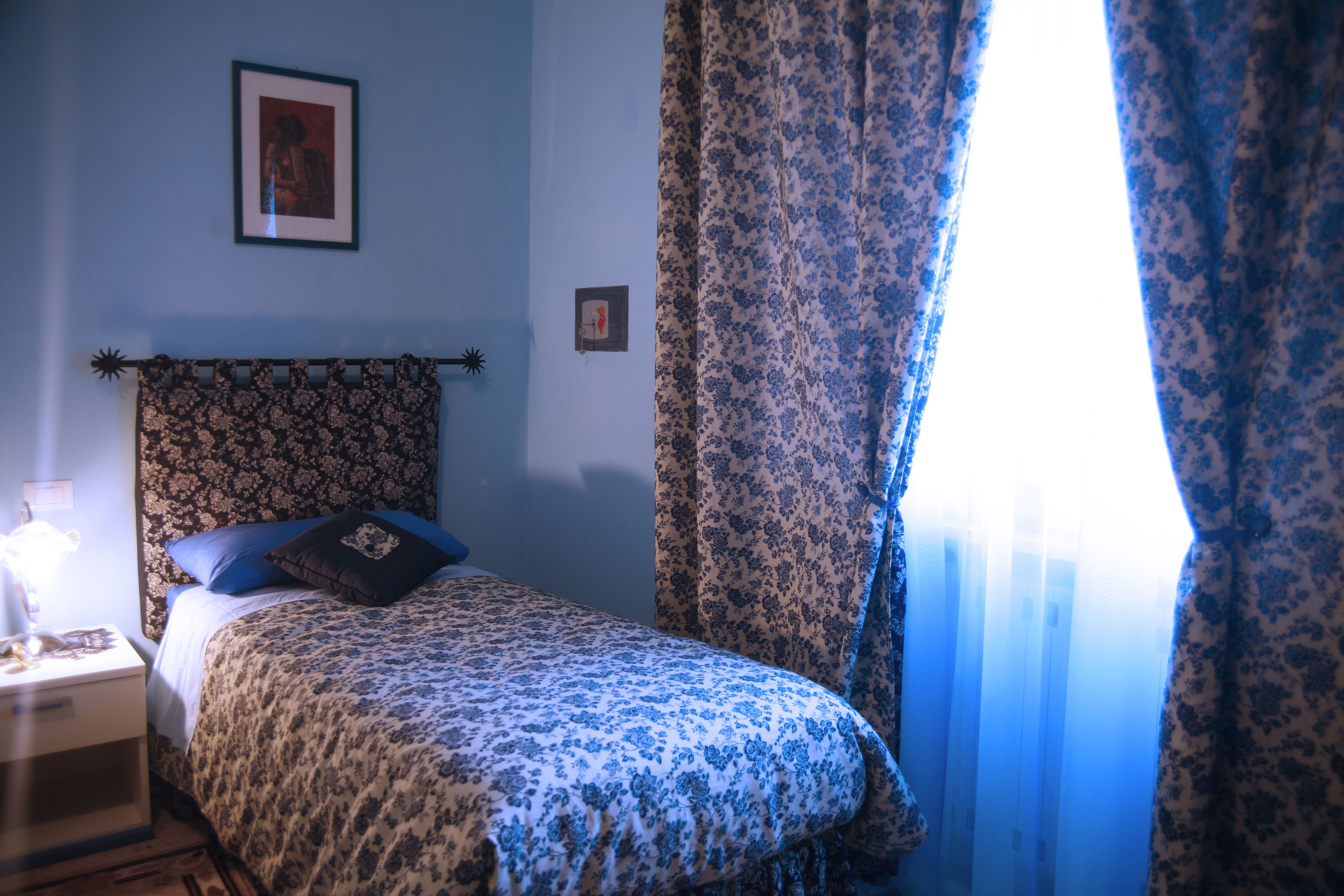 Un dormitori amb un llençol estampat, una tauleta de nit amb una làmpada i una finestra amb cortines florals.