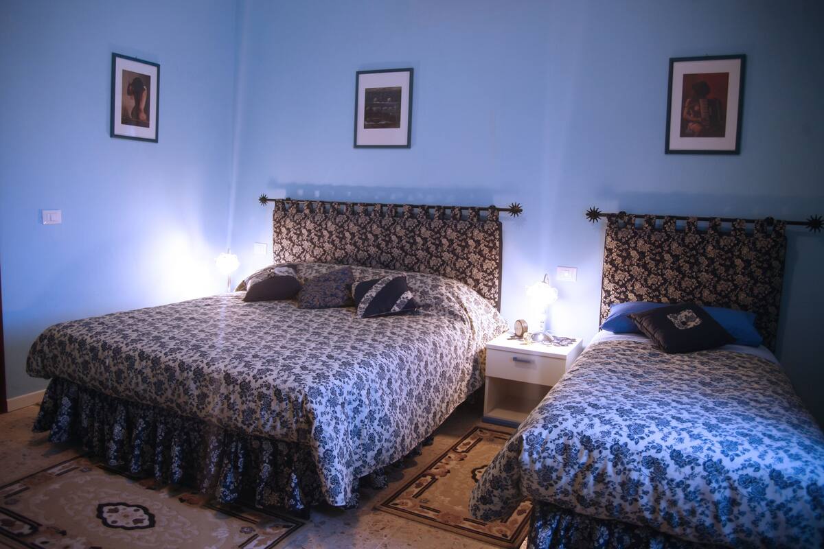 A room with two beds, each with a floral-patterned bedspread and matching headboards. There are bedside tables with lamps and framed pictures on the walls.