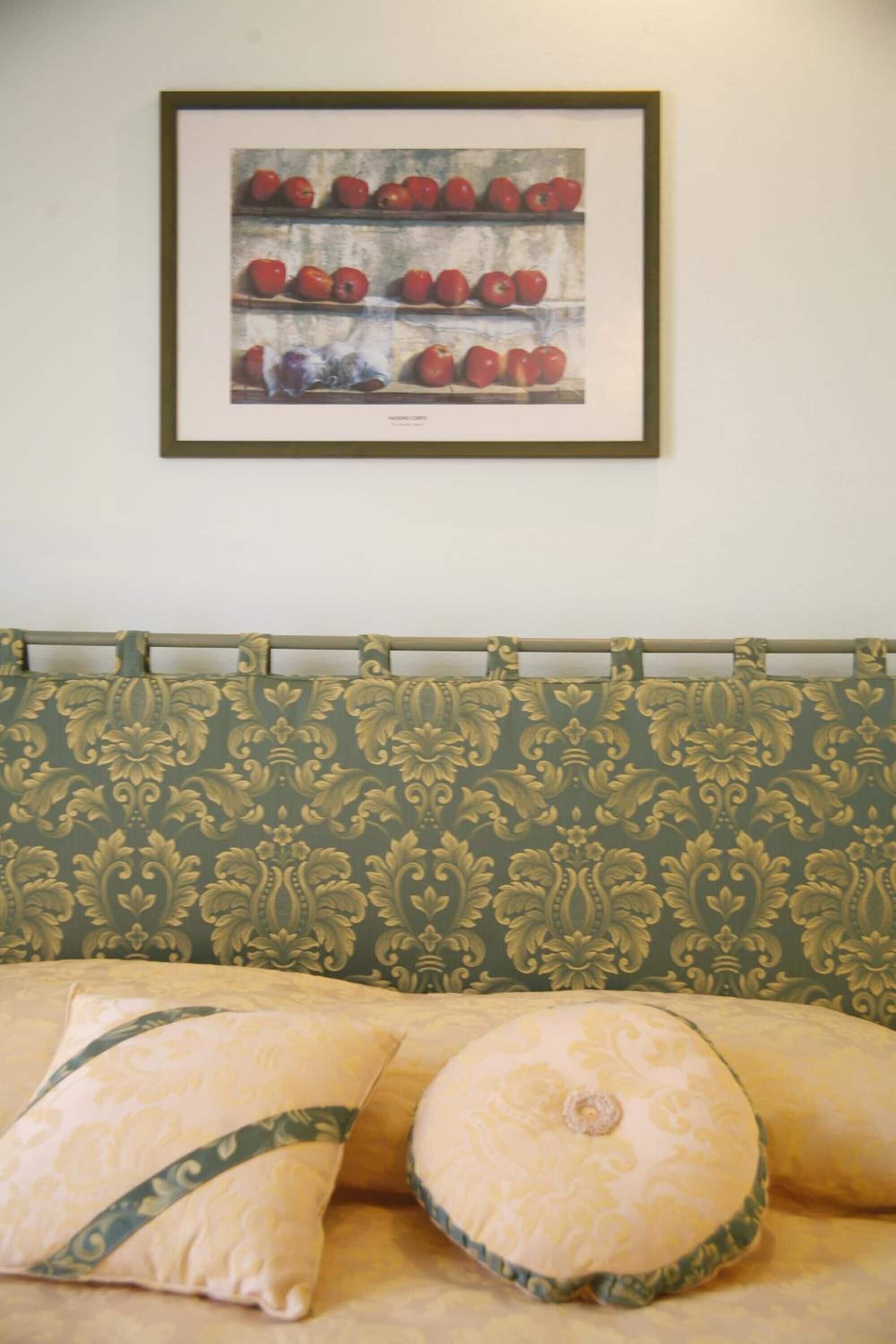A bedroom with a patterned green and beige headboard, a framed picture of apples on the wall, and two pillows with a similar pattern.