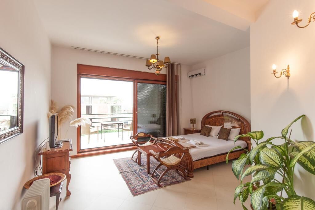 A bedroom with a bed, a desk, and a chair, a large window, and a plant.