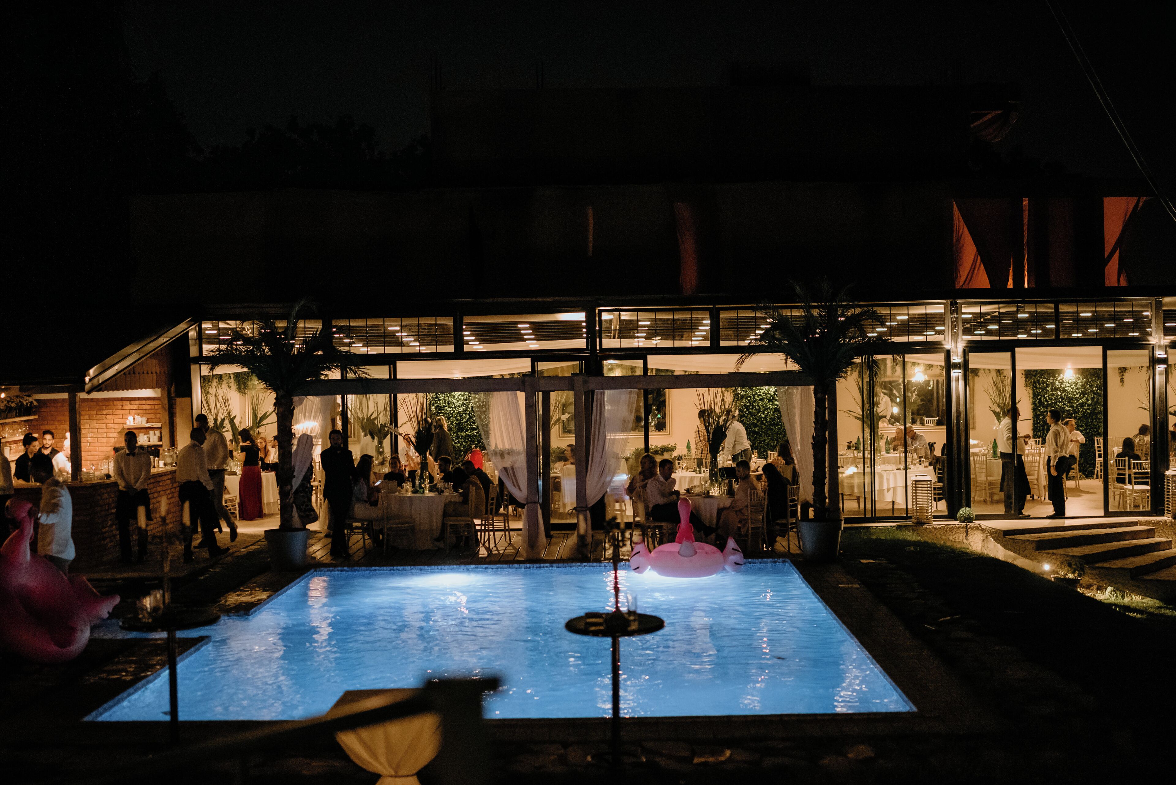 A pool area with a glass enclosure, a flamingo float, and people dining inside.