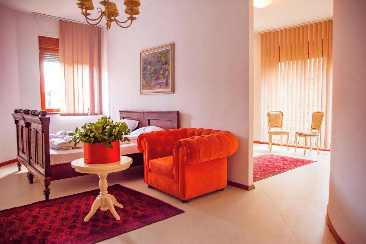 Une pièce avec un lit, un fauteuil rouge, une petite table avec une plante et un tableau au mur.