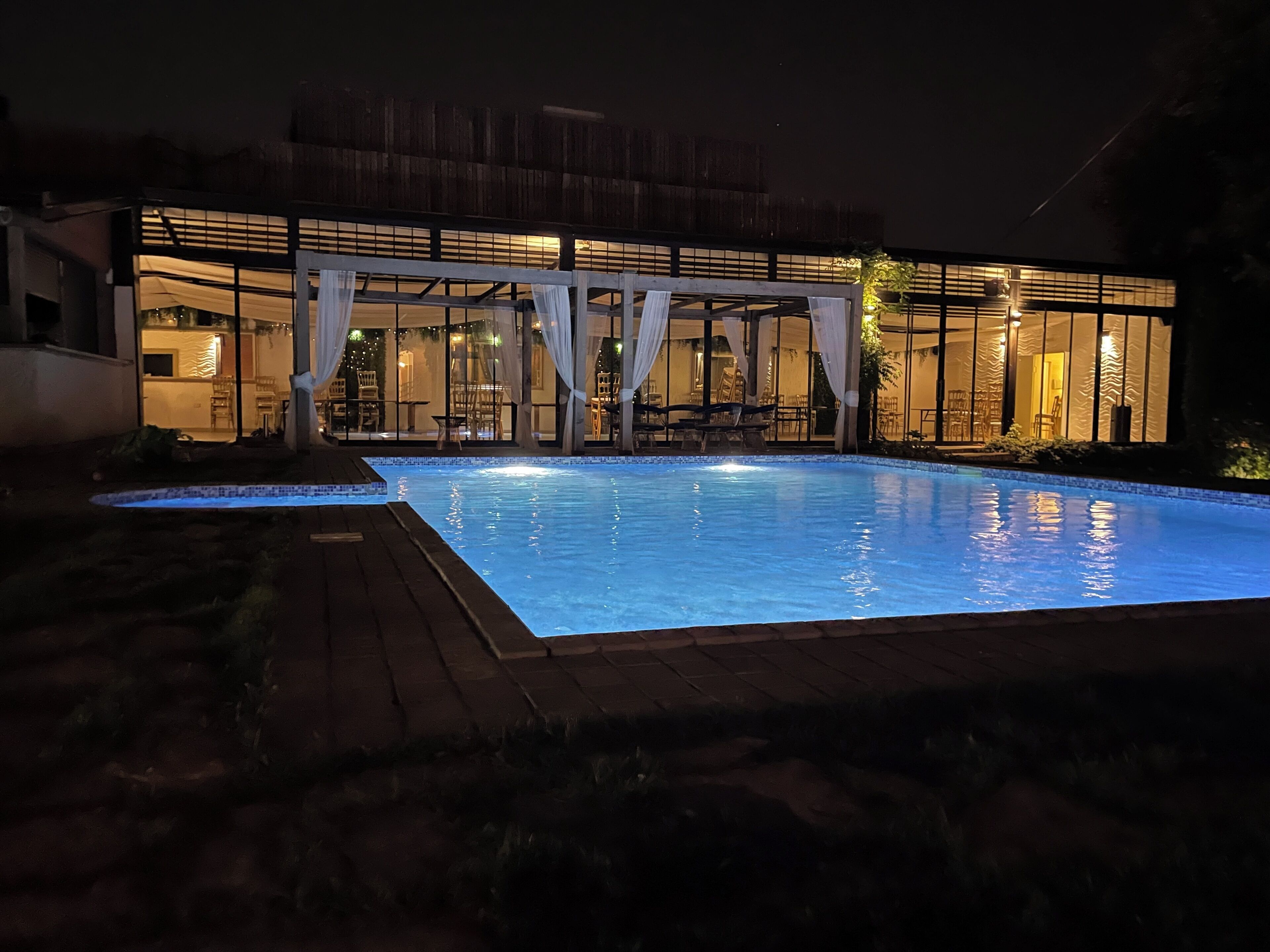 Una piscina con plataforma de madera, rodeada por un espacio exterior cubierto con sillas y mesas.