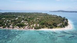 An aerial view of a tropical island with clear turquoise waters, surrounded by lush greenery and small buildings. - Sandy Beach Bungalows (Gili Air)