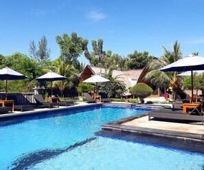 Una piscina con lettini e ombrelloni, circondata da vegetazione tropicale e edifici.
