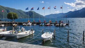 A marina with boats docked, a dock with people, and mountains in the background. - Red View Apartment with Balcony Terrace, Ronco sopra Ascona, Switzerland (Ronco sopra Ascona)
