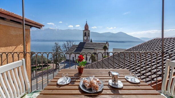 A balcony with a table set for two, overlooking a town with a church and mountains. - Red View Apartment with Balcony Terrace, Ronco sopra Ascona, Switzerland (Ronco sopra Ascona)