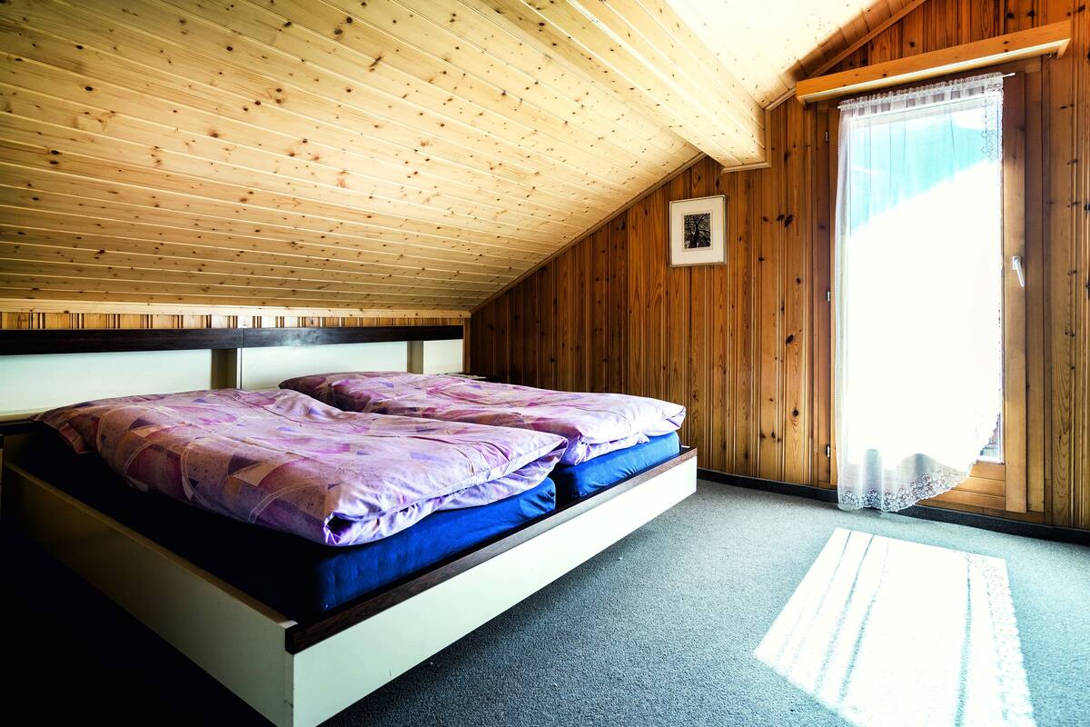 A bedroom with a sloped ceiling, wooden walls, a bed with purple and blue bedding, and a window with a sheer curtain.
