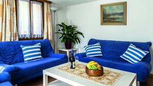 Una sala de estar con dos sofás azules, una mesa de centro blanca con un frutero, una botella de vino y una planta en maceta.