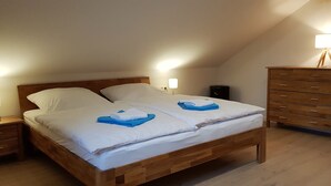 Uma cama de solteiro com estrutura de madeira e roupa de cama branca, uma mesa de cabeceira com abajur e uma cômoda de madeira com gavetas.