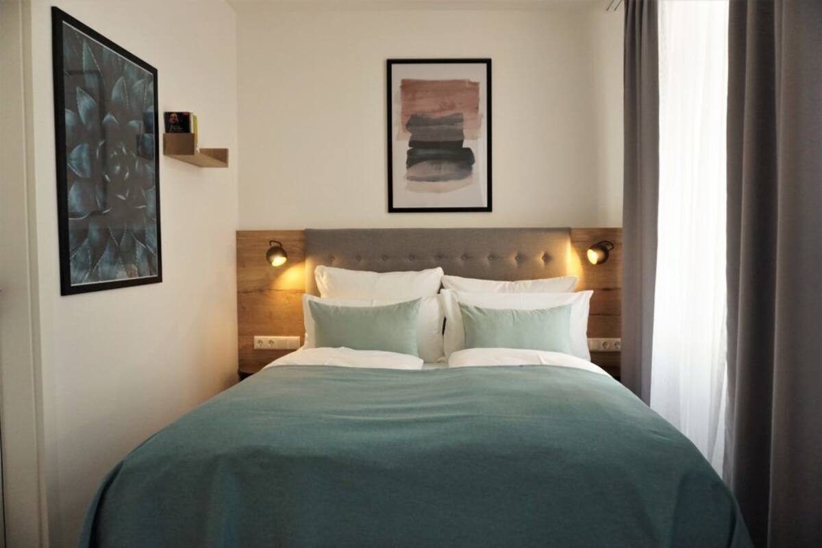 A neatly made bed with a wooden headboard, two wall-mounted reading lights, and a framed abstract painting above it.