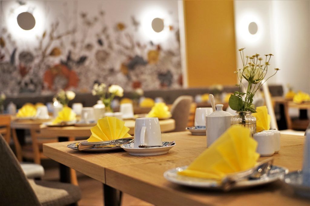 Un comedor con mesas de madera dispuestas con tazas blancas, platos y servilletas amarillas.