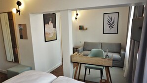 A modern hotel room with a bed, a desk with a chair, a sofa, and a small table. There are framed pictures on the walls and a lamp on the desk. - dieSonne (Weimar)