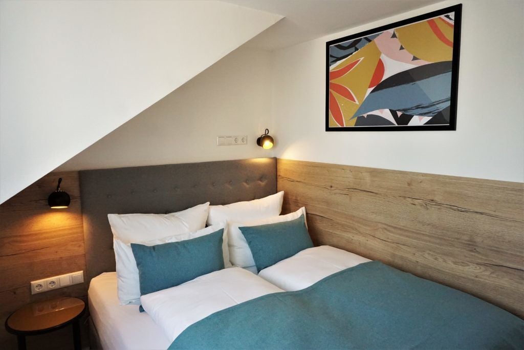 Un dormitorio con cabecera de madera, cama con ropa de cama blanca y turquesa, una mesita de noche pequeña y una l ámpara de pared.