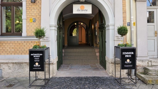 An entrance to a building with a sign reading "de Sonne" above the door. There are two signs on either side of the entrance, one for a buffet and the other for a room with a view. - dieSonne (Weimar)