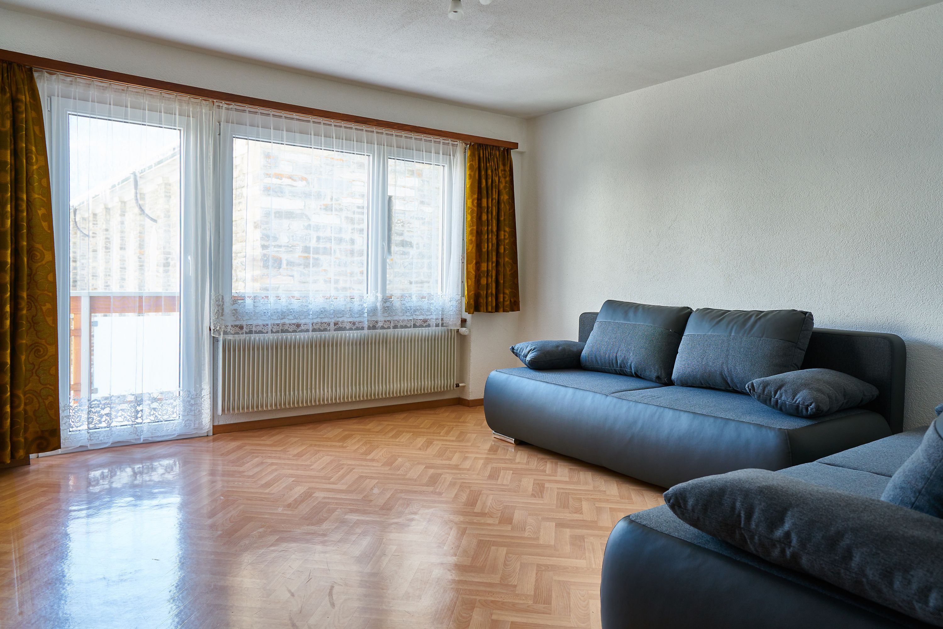 A modern living room with a wooden parquet floor, a blue sofa, and a large window with curtains.