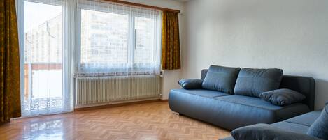A modern living room with a wooden parquet floor, a blue sofa, and a large window with curtains.