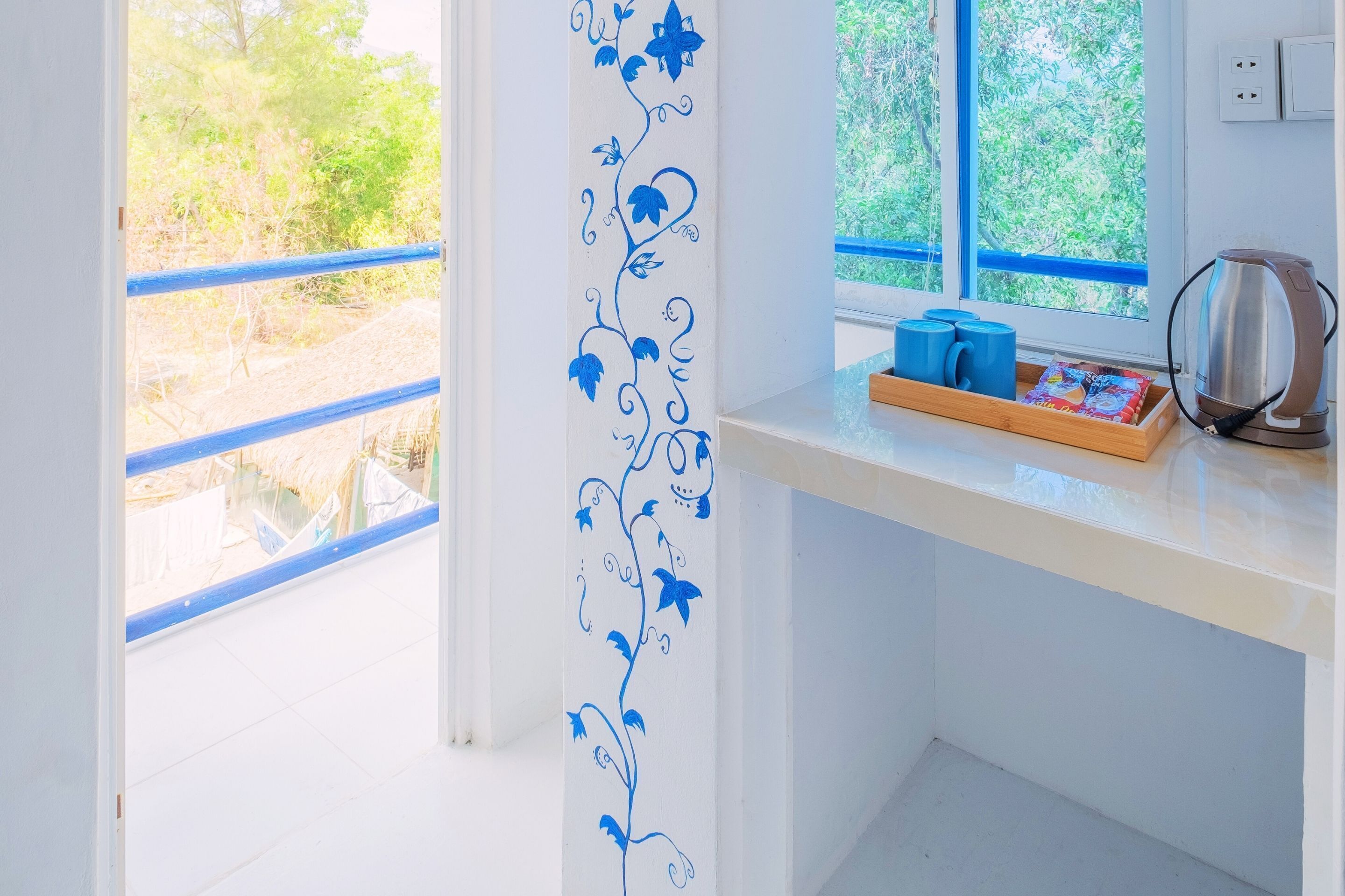 Una habitación luminosa con una pared blanca y una ventana con barandilla azul. Hay una estantería con una tetera, tazas y una caja de pañuelos.