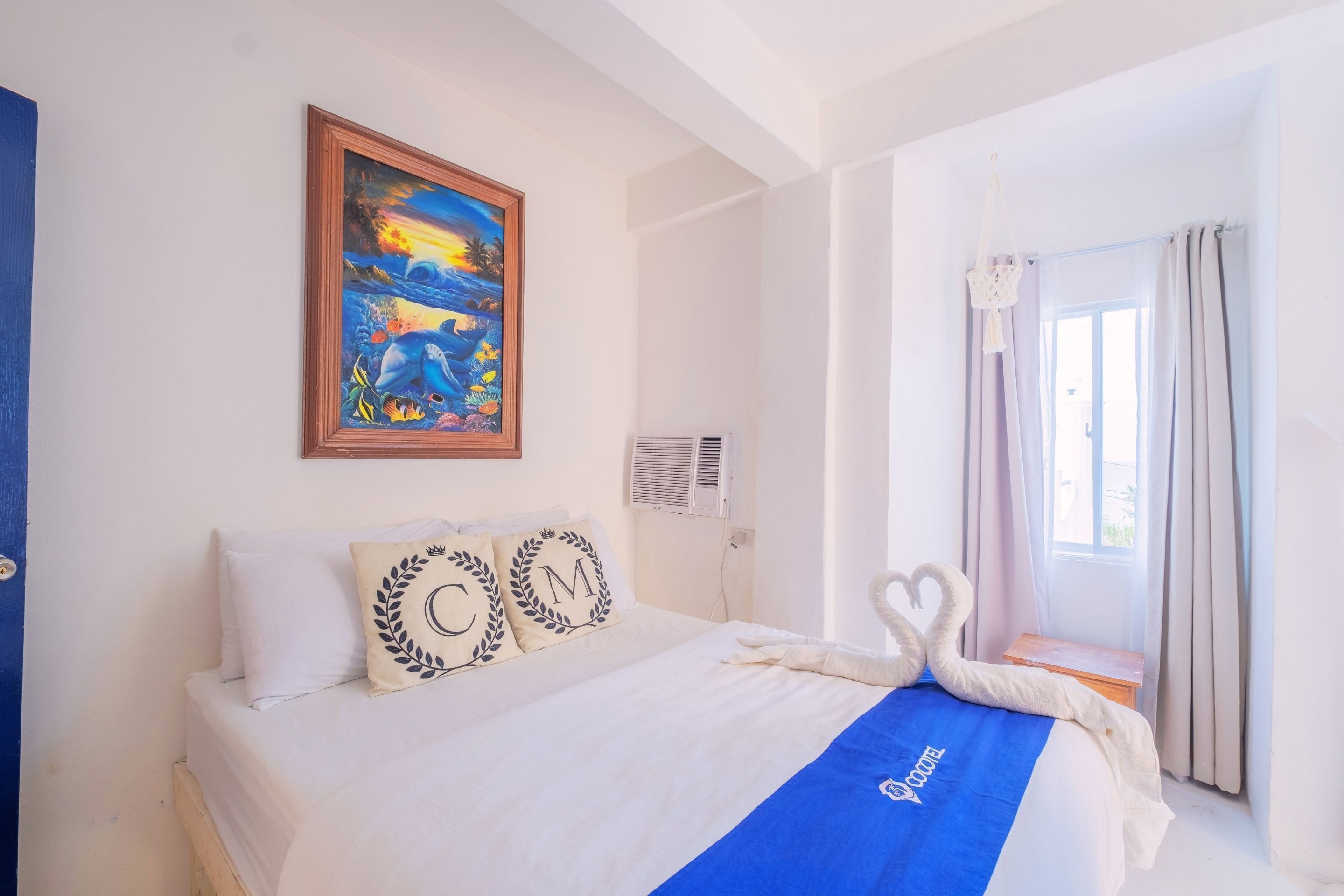 A hotel room with a bed, a painting of a tropical scene, a window with curtains, and a decorative swan on the bedspread.