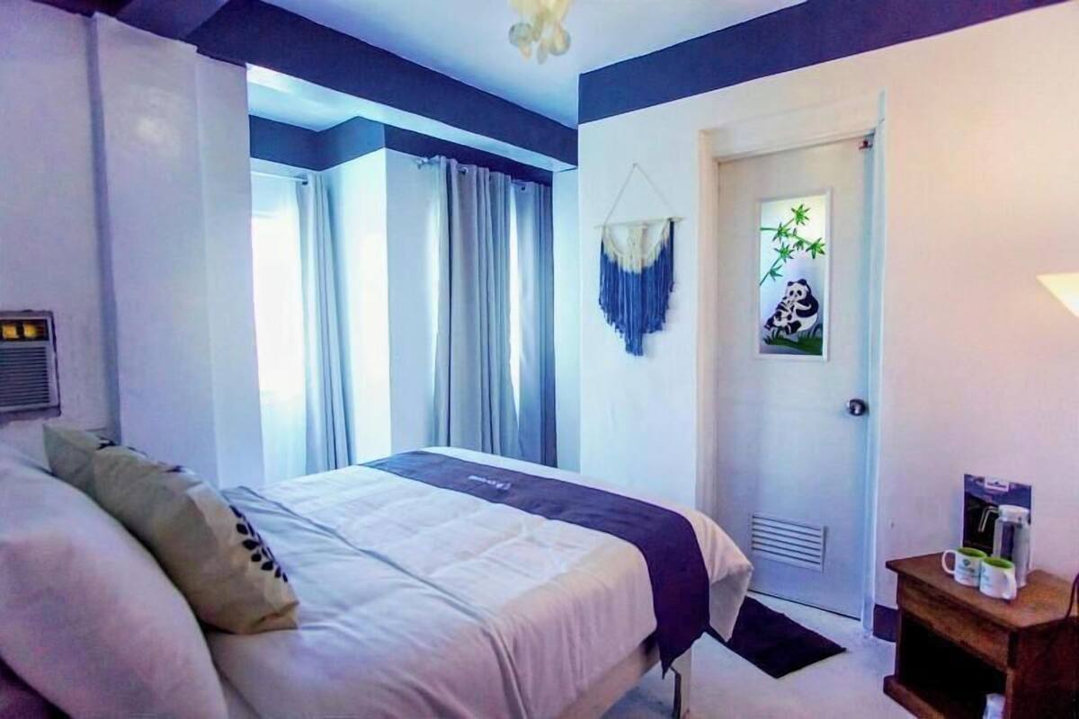 Une chambre d’hôtel avec un lit, des rideaux, une porte, un élément de décoration murale et une petite table sur laquelle sont posés divers objets.