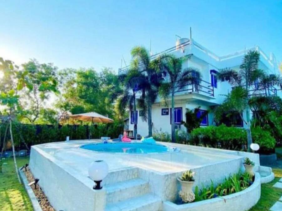 A modern two-story house with a swimming pool, surrounded by lush greenery and palm trees.