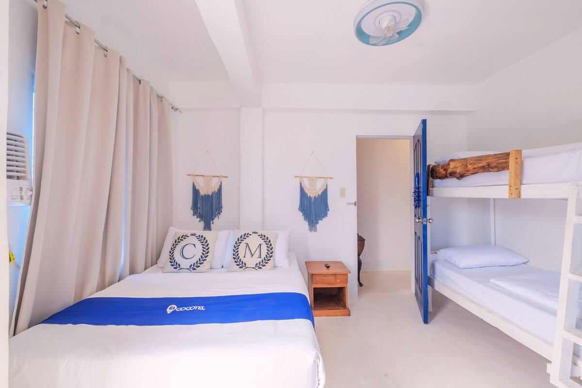 Une chambre d’hôtel avec un lit simple, un lit superposé, une petite table de chevet en bois et un ventilateur de plafond.