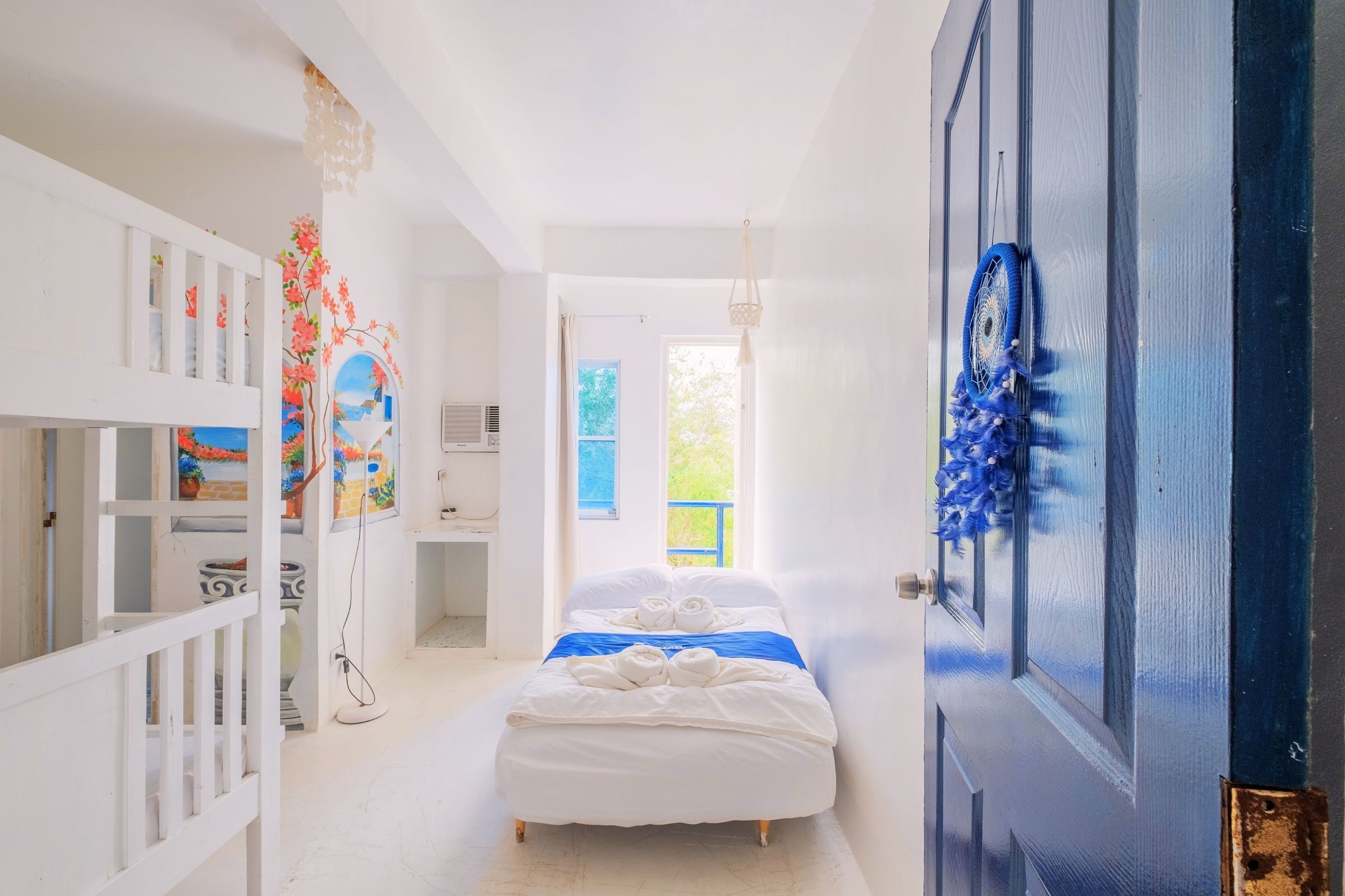 Una habitación luminosa y blanca con una cama, una puerta adornada con un adorno azul, y vistas a la vegetación exterior.