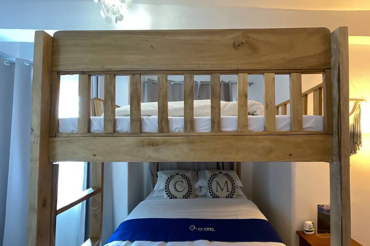 Un lit superposé avec une tête de lit en bois, deux oreillers ornés d’initiales, une couvre-lit bleue et une table de chevet avec une tasse.