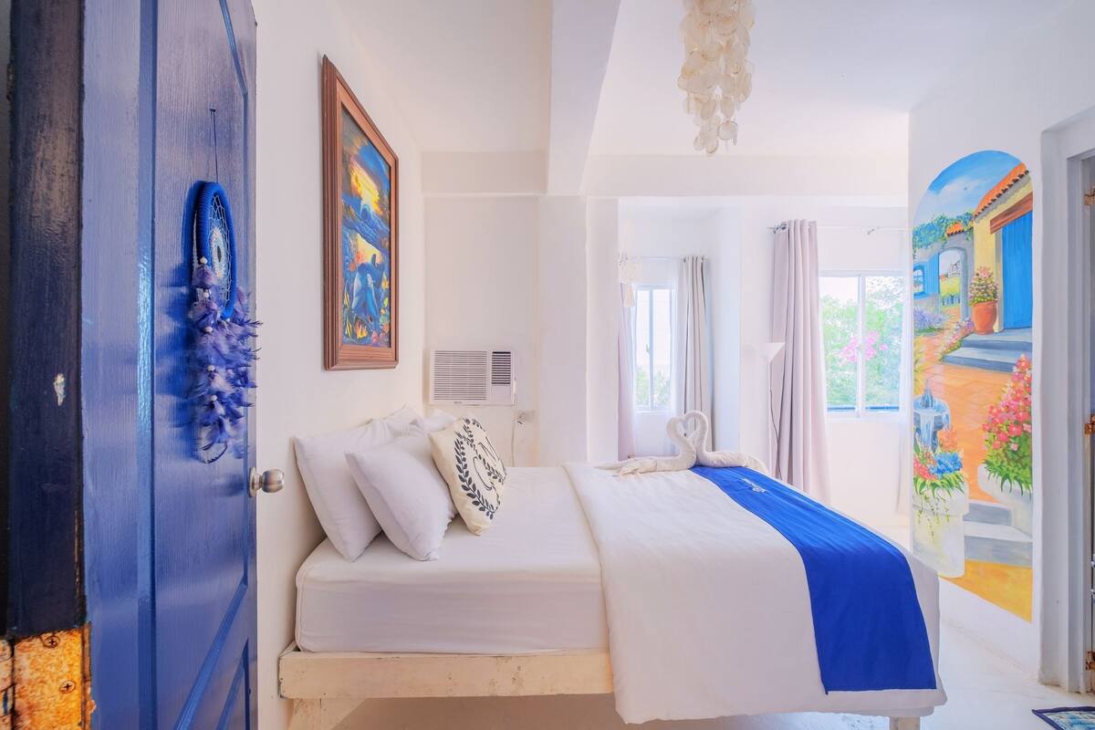 Une chambre à coucher avec un lit, un tableau au mur, un luminaire au plafond et une vue sur l’extérieur.