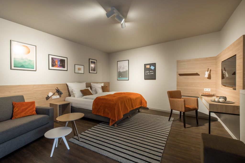 A modern hotel room with a bed, a sofa, a desk, and a chair. There are framed pictures on the wall and a window with a view.