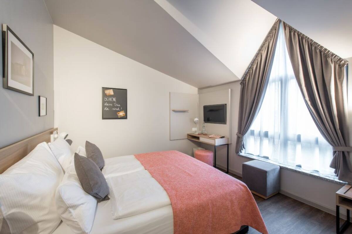 A modern hotel room with a bed, a desk, a TV, and a window with curtains.