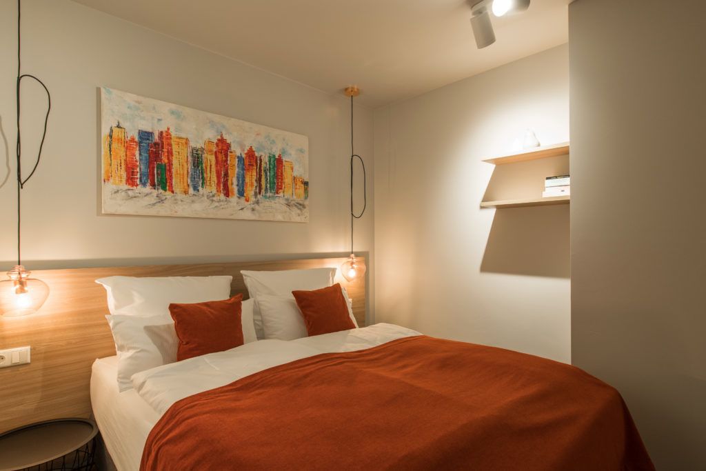 A modern hotel room with a bed, a cityscape painting, and a bedside table with a lamp.