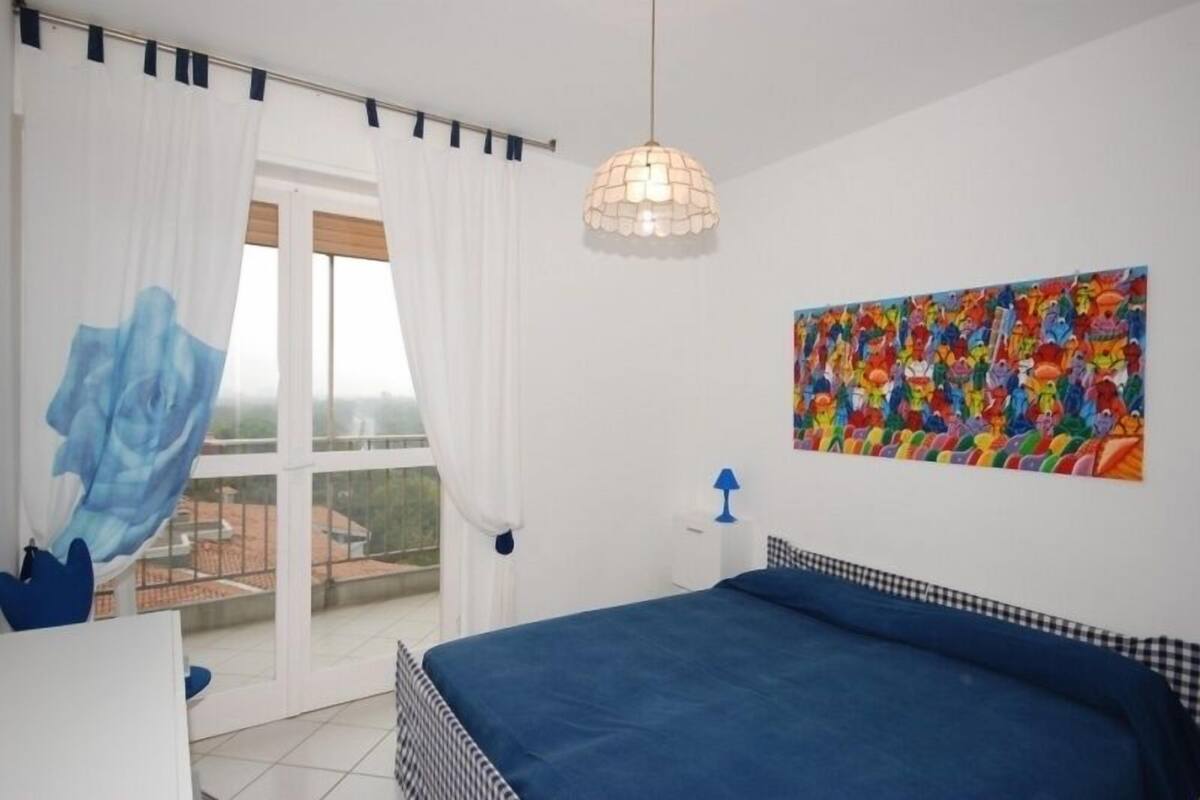 A bedroom with a bed, a window with curtains, a hanging light fixture, and a colorful painting on the wall.