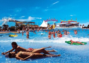 A large outdoor swimming pool with multiple lanes, a water slide, and a ship named 'Culturallandia Aquarium' in the background. - Alideco (Lignano Sabbiadoro)