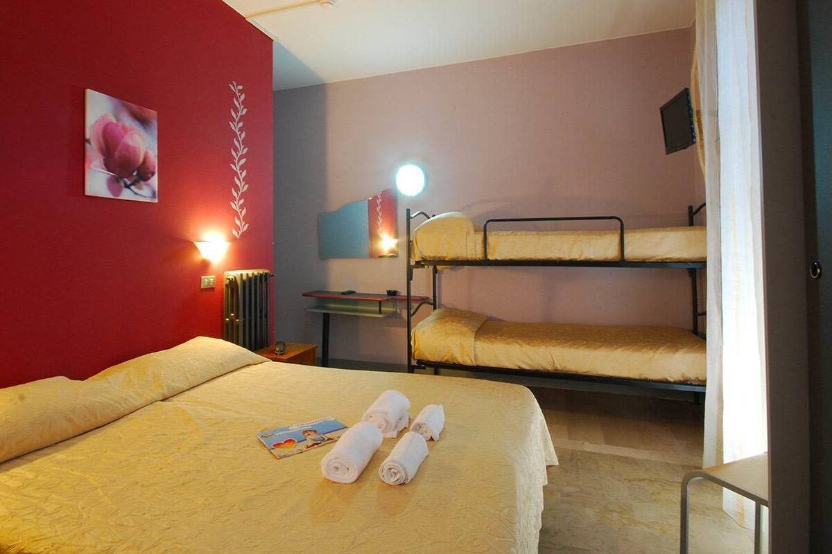 Quarto de hotel com uma cama grande, duas beliches, uma mesa de cabeceira com abajur e uma parede vermelha com arte floral.