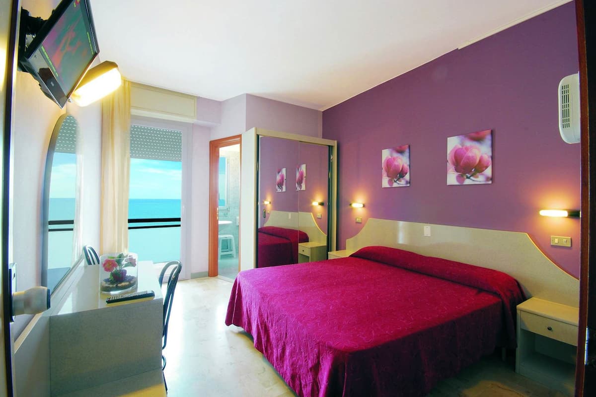 Quarto de hotel com cama, mesas de cabeceira, escrivaninha e uma cadeira. Tem uma janela com vista para o mar.