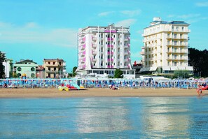 A beachfront hotel with multiple floors, balconies, and a beach area with umbrellas and sun loungers. - Hotel Kent (Riccione)