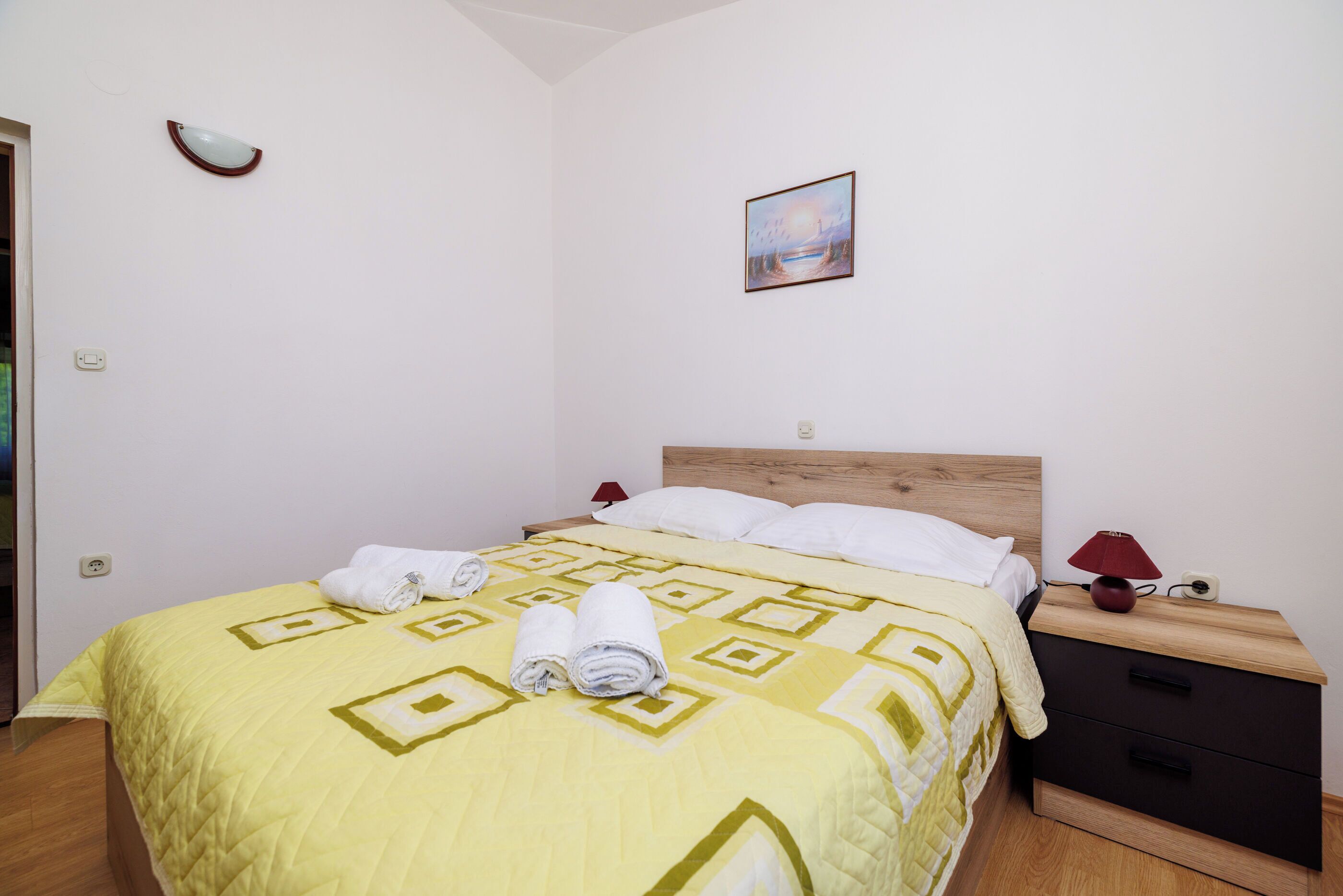 Ein ordentlich bezogenes Bett mit einer gelb-weißen, gemusterten Tagesdecke, einem hölzernen Kopfteil und einem Nachttisch mit Lampe und Bilderrahmen.