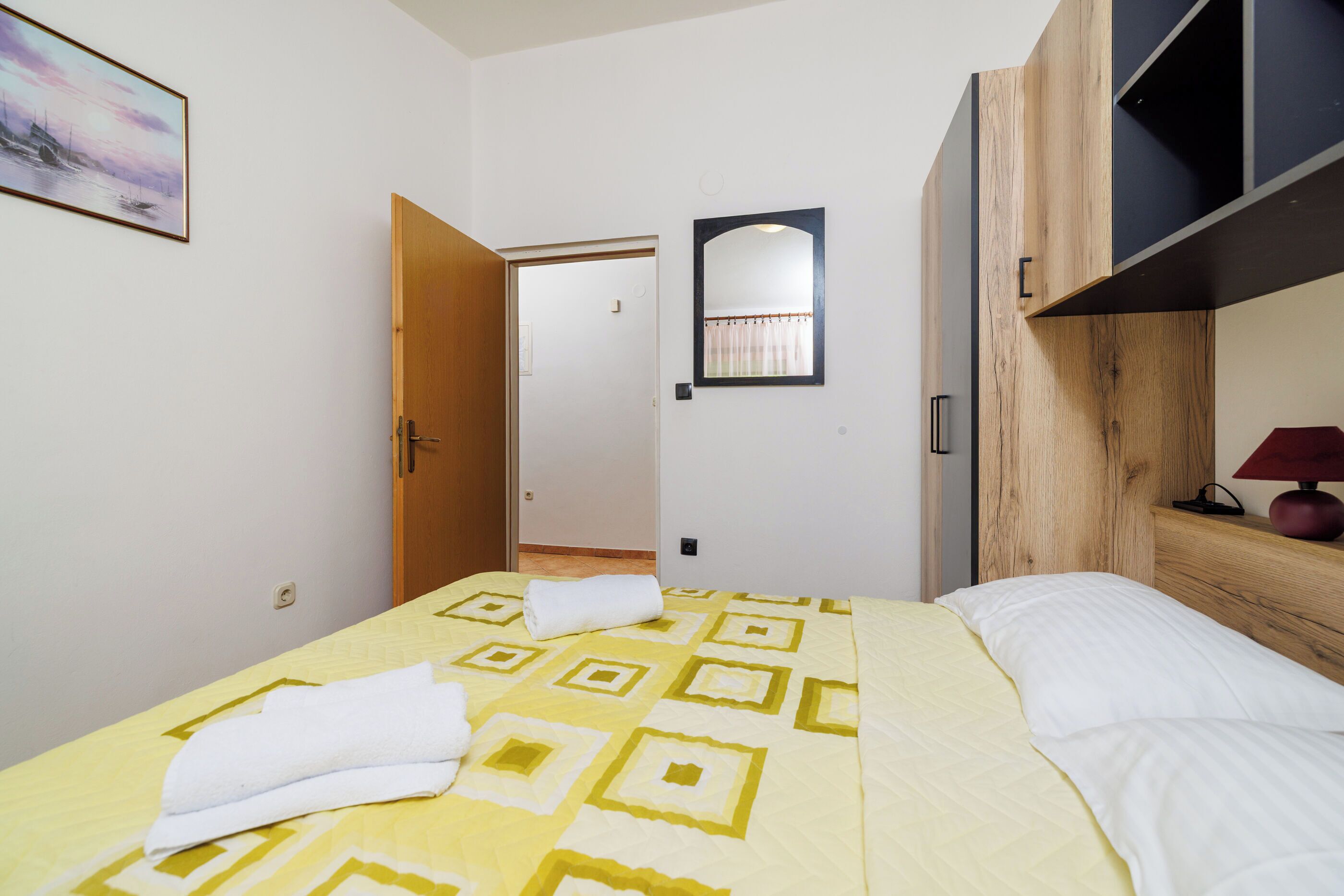A hotel room with a bed, a wooden wardrobe, a mirror, and a painting on the wall.