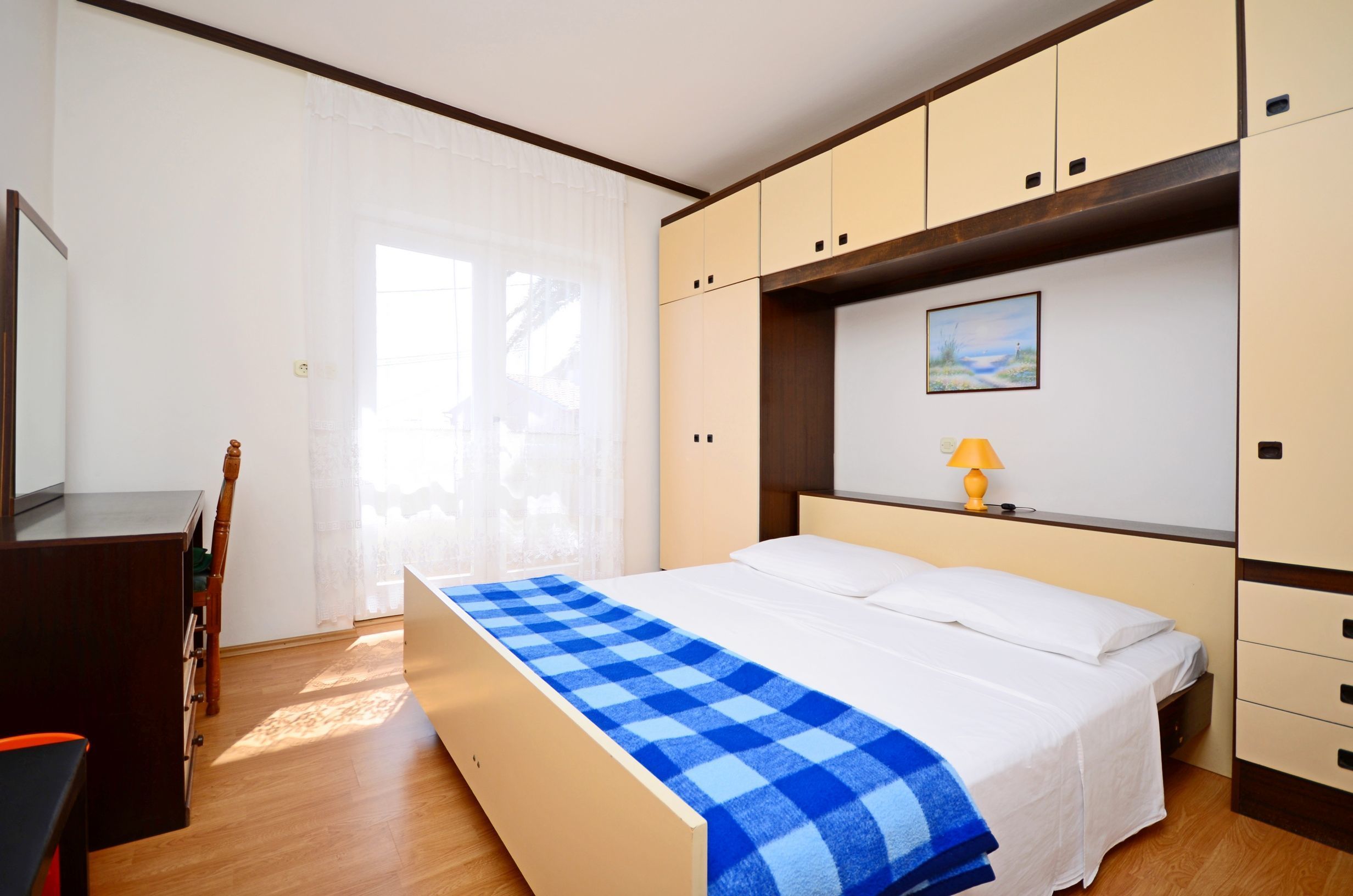 Una camera da letto con un letto, una scrivania e una sedia. Si vede una finestra con tende, un quadro al muro e una lampada sulla scrivania.